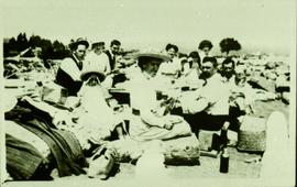 eight adults and three children picnicing on a beach. On the right side of the picture, Phillip Francis Hayward is holding infant daughter, Frances, and Ella is sat next to him