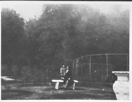 Minnie Hayward and daughter on a bench in the rose garden, tennis court behind.