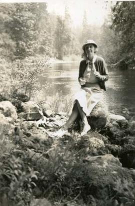 Muriel with drink and cigarette on a rock beside a river